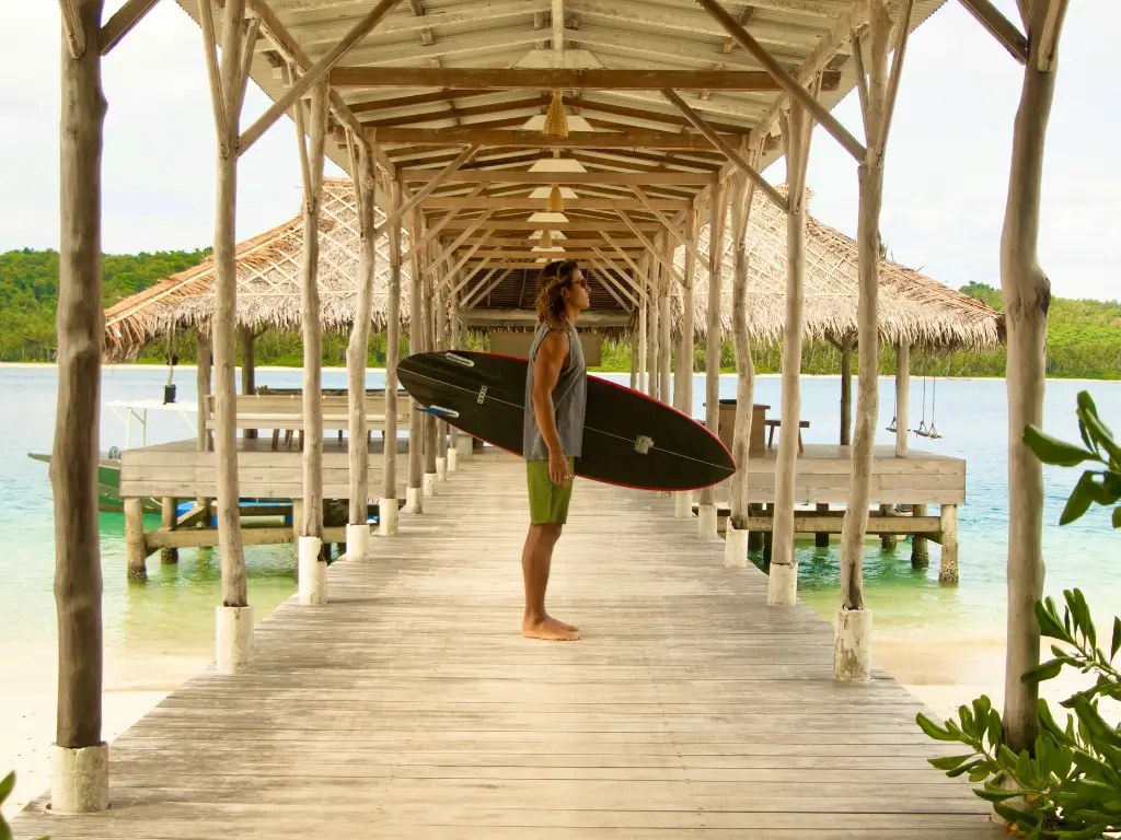 A man is standing n the board walk with a board in his hands - mentawai surf resort